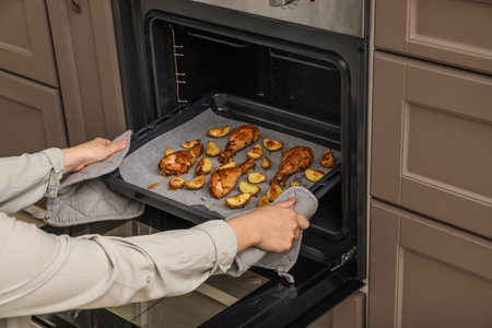 Woman taking baking tray with paper, chicken legs and potato from oven, closeupの写真素材