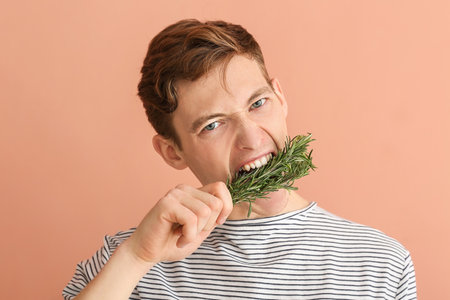 Angry young man eating fresh rosemary on color background