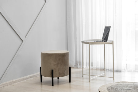 Interior of stylish living room with light curtains, pouf, and table with laptop