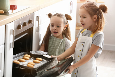 Cute little sisters taking tasty cookies from oven in kitchenの写真素材