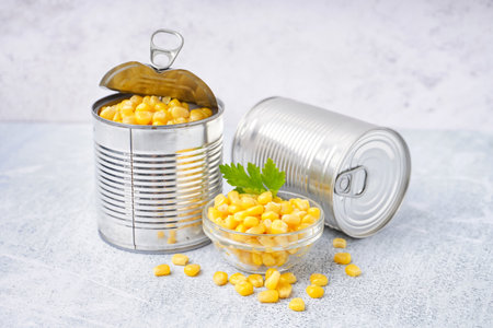 Tin cans, bowl with corn kernels and parsley leaf on gray tableの写真素材