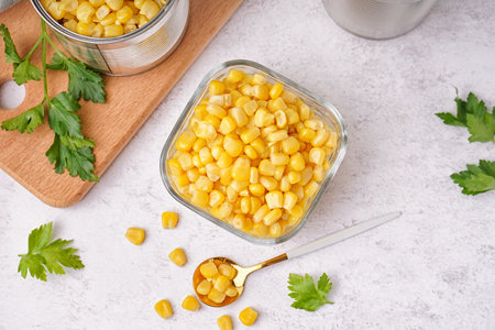 Bowl with corn kernels, cutting board, spoon and parsley on white table