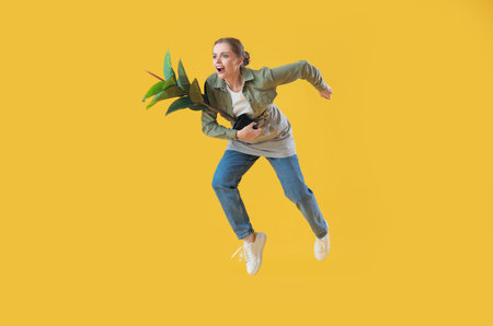 Female gardener with houseplant jumping on yellow background