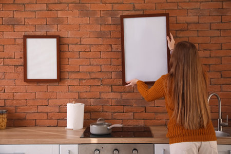 Young woman hanging blank frame on brick wall in kitchen, back viewの写真素材