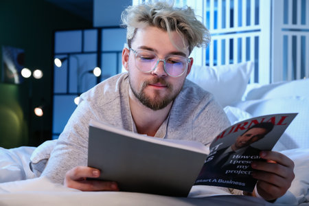 Young man wearing glasses while reading magazine at nightの写真素材