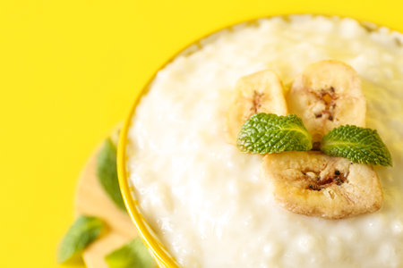 Bowl with delicious rice pudding, banana slices and mint leaves on yellow background, closeupの写真素材