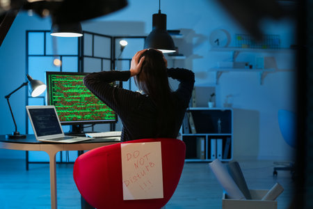 Female programmer working with computer and paper sheet with text DO NOT DISTURB in office at nightの写真素材