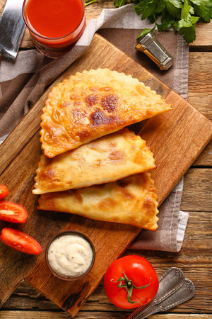 Cutting board with tasty chebureks, sauce and tomato juice on wooden background