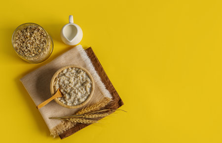 Bowl with oatmeal, flakes in jar, pitcher and wheat ears on yellow background