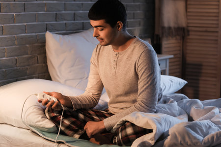 Young man sitting on electric heating pad in bedroom at nightの写真素材