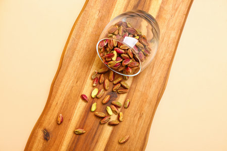Board with overturned glass of tasty pistachio kernels on beige background