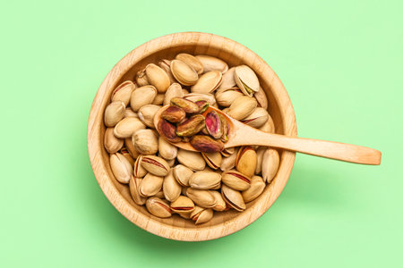 Bowl with tasty pistachio nuts and spoon on green background