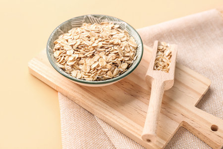 Wooden board with bowl and scoop of raw oatmeal on color background, closeup