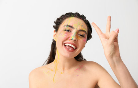 Beautiful young woman with paint on her face showing victory gesture against light background