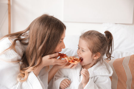 Young woman with her little daughter eating pizza in the bedroomの写真素材