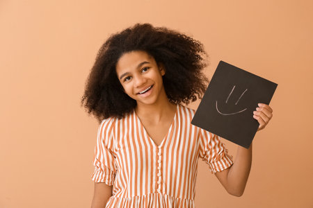 African-American teenage girl holding chalkboard with drawn emoticon on color backgroundの写真素材