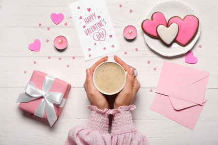 Woman with cup of latte, text HAPPY VALENTINE'S DAY, gift and cookies on white wooden backgroundの写真素材
