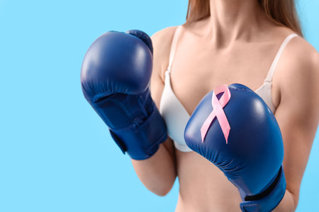 Young woman in underwear with pink ribbon and boxing gloves on blue background. breast cancer awareness conceptの写真素材