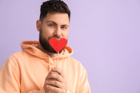 Young bearded man with paper heart for Valentine's Day on lilac background, closeupの写真素材