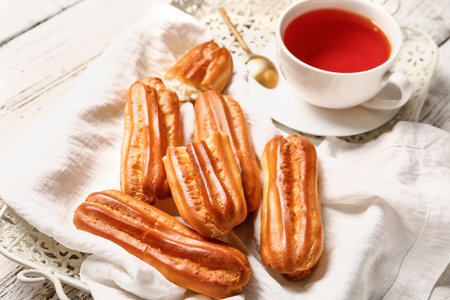 Tray with tasty eclairs and cup of tea on light wooden background, closeup