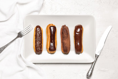 Plate with tasty chocolate eclairs and cutlery on light backgroundの写真素材