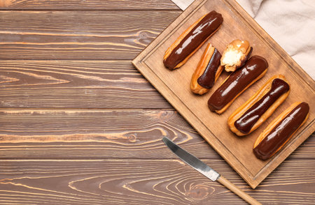 Cutting board with tasty chocolate eclairs on wooden background