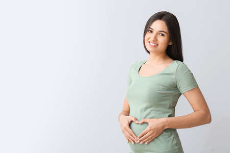 Beautiful pregnant woman making heart with her hands on light background