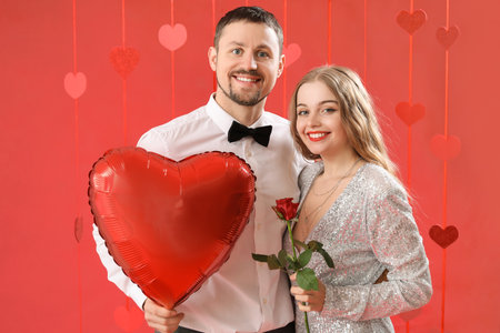 Happy young couple with heart-shaped balloon and rose on red background. Valentine's Day celebrationの写真素材