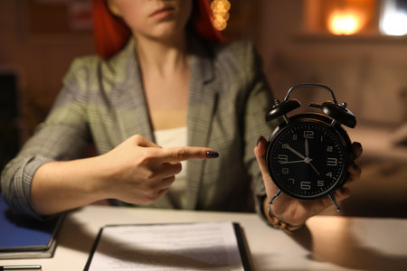 Young woman pointing at alarm clock in office at night