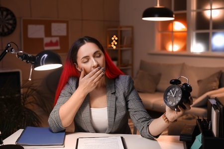 Tired young woman with alarm clock in office at nightの写真素材