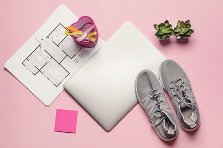 Laptop with drawings, office supplies and sports shoes on color background