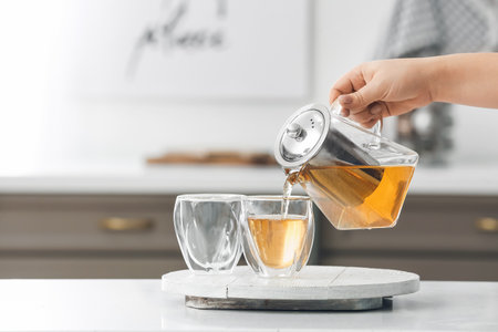 Woman pouring hot tea from teapot into glasses in kitchenの写真素材