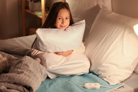 Little girl with pillow sitting on electric heating pad in bedroom at nightの写真素材