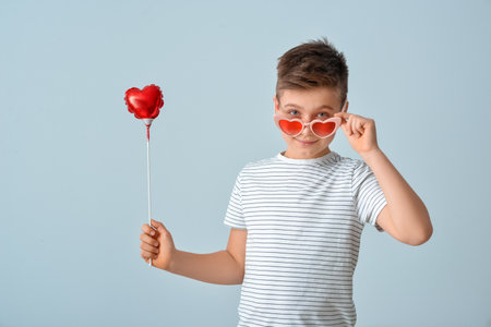 Cute little boy in stylish heart-shaped sunglasses and with air balloon on gray backgroundの写真素材