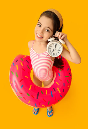 Cute little girl in swimsuit, with alarm clock and inflatable ring on color background