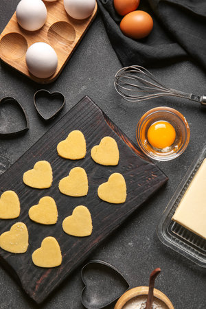 Wooden board with raw heart shaped cookies, ingredients and cutters on dark background. valentine's...