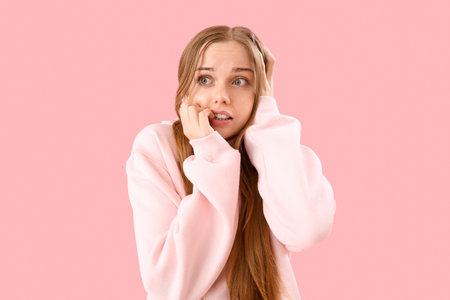 Young woman in hoodie biting nails on pink backgroundの写真素材