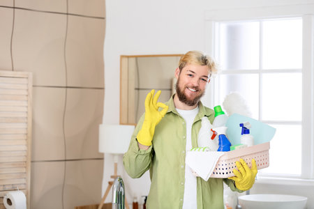 Young man with cleaning supplies showing OK in bathroomの写真素材