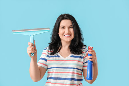 Mature woman with detergent and squeegee on color background