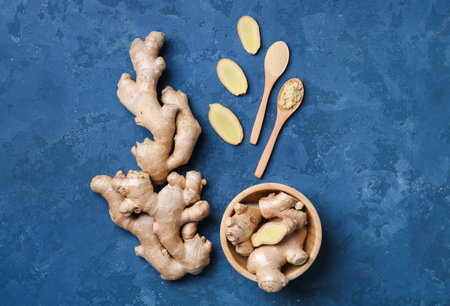 Wooden plate with fresh ginger roots, slices and spoons of powder on blue grunge background