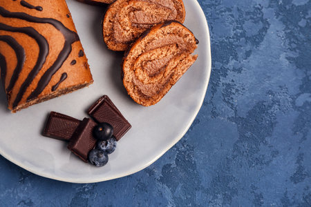 Plate with pieces of delicious sponge cake roll, chocolate and blueberry on blue tableの写真素材