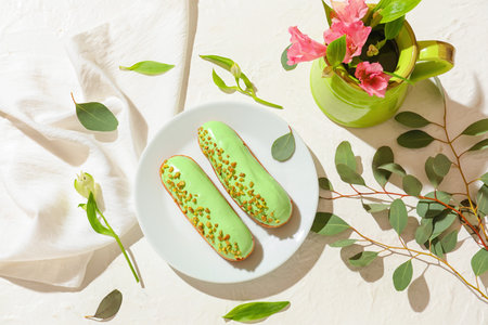 Plate with delicious eclairs and jug of flowers on light background