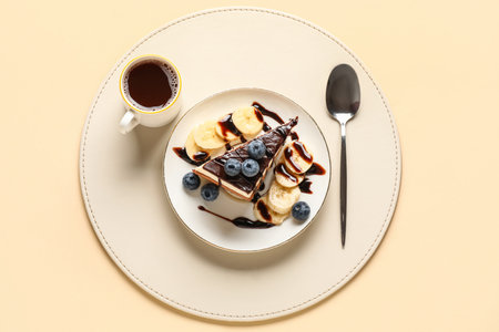 Table setting with piece of birds milk cake, blueberries, chocolate topping, banana slices, cup of coffee and spoon on beige backgroundの写真素材