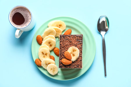 Table setting with piece of birds milk cake, almond nuts, banana slices, cup of coffee and spoon on blue backgroundの写真素材
