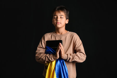 Little boy with Holy Bible and flag of Ukraine praying on black backgroundの写真素材