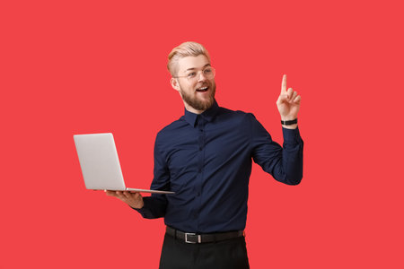Young man with laptop pointing at something on color backgroundの写真素材