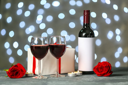 Glasses of wine, bottle, burning candles, gift and rose flowers on gray table against blurred lights. Valentine's Day celebrationの写真素材