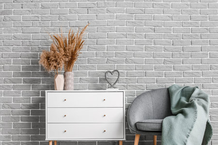 Modern chest of drawers with vases and armchair near brick wall in roomの写真素材