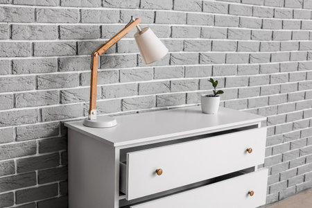 Chest of drawers with lamp and houseplant on brick background