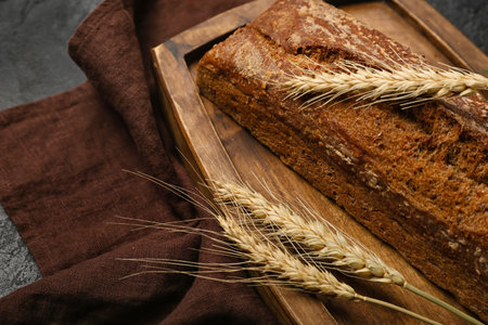 Board with loaf of fresh rye bread and wheat ears on black background